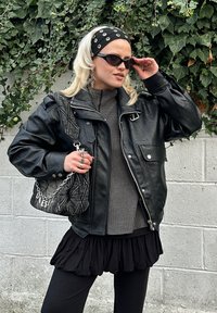 Čierna kožená bunda s vysokým golierom, sivý rebrovaný sveter, čierna vrstvená sukňa a vzorovaná kabelka. Doplnky zahŕňajú slnečné okuliare a kruhové náušnice.