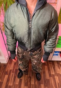 Zöld bombázó dzseki cipzáros elülső résszel, nagy kapucnival és bordázott mandzsettákkal. Álcázott zsebes nadrággal és fekete fűzős bakancsokkal viselve.