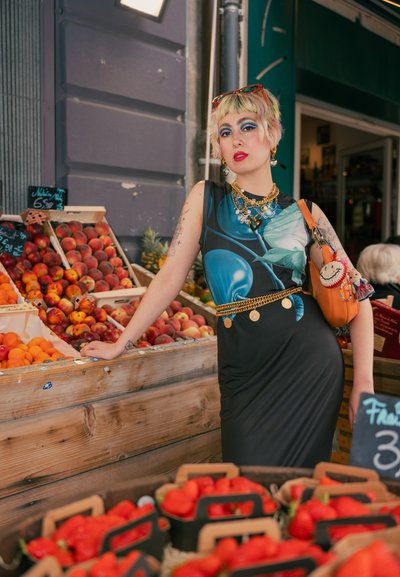 Vestido oscuro con estampado floral turquesa, acentuado por un cinturón de cadena dorada y joyas. Bolso naranja con llavero de peluche. Fondo de mercado de frutas.