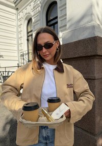 Giacca in velluto a coste beige con collo marrone, t-shirt bianca, due tazze di caffè in un portabicchieri di cartone e un tubetto dorato in mano; ambiente esterno.