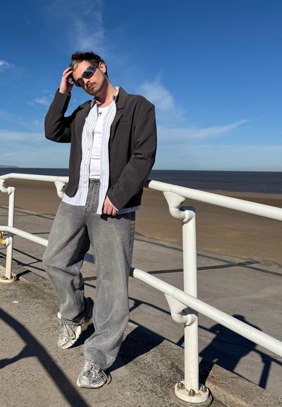 Chaqueta gris sobre una camisa de botones a rayas y una camiseta blanca, combinada con jeans grises holgados y zapatillas con estampado. De pie junto a una barandilla.
