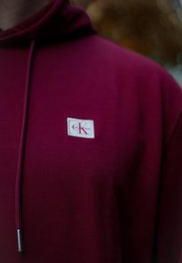 Burgundi kapucnis pulóver zsinórral, fehér címkével, amelyen a "Calvin Klein" felirat és egy stilizált piros "K" található. Puha, texturált anyag.