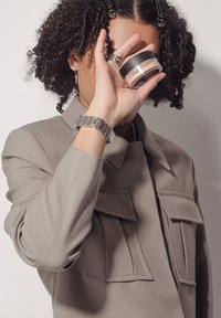 Veste beige avec deux poches avant, montre argentée au poignet et personne tenant un bocal transparent avec un couvercle noir et doré devant son visage.