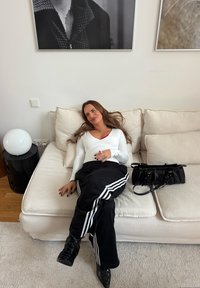 Une femme portant un haut à manches longues blanc et un pantalon de survêtement noir avec des bandes blanches est assise sur un canapé clair, à côté d'un sac à main noir et d'une lampe sphérique.