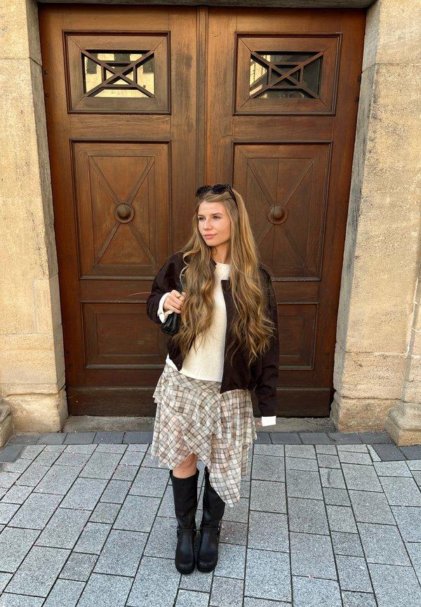 Jeune femme aux longs cheveux blonds, portant une jupe à carreaux, des bottes noires et une veste foncée, debout sur un pavé de pierre devant de grandes portes en bois.