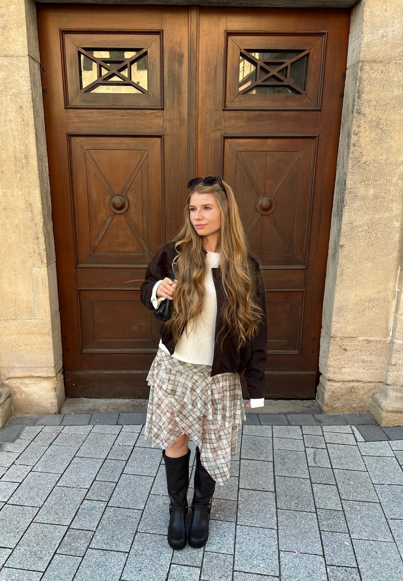 Jeune femme aux longs cheveux blonds, portant une jupe à carreaux, des bottes noires et une veste foncée, debout sur un pavé de pierre devant de grandes portes en bois.