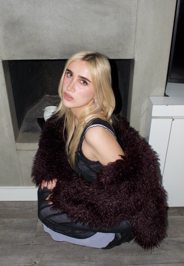 Blonde vrouw in een zwarte jurk en een donker, pluizig jasje hurkt binnen op een houten vloer, kijkt met een neutrale uitdrukking omhoog bij een betonnen haard.