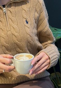 Ruce držící bílý keramický hrníček s latte artem, oblečené v béžovém svetr s pleteným vzorem a s diamantovým prstenem na levé ruce.