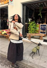 Femme aux cheveux bouclés ajuste son col, tenant une laisse pour un petit chien en veste verte devant un café avec un stand de fruits à proximité.