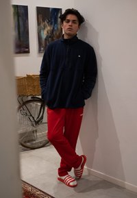Marine fleece pullover with a half-zip, teamed with vibrant red tracksuit bottoms. White sandals boast red stripes. The background includes a bicycle and wall art.