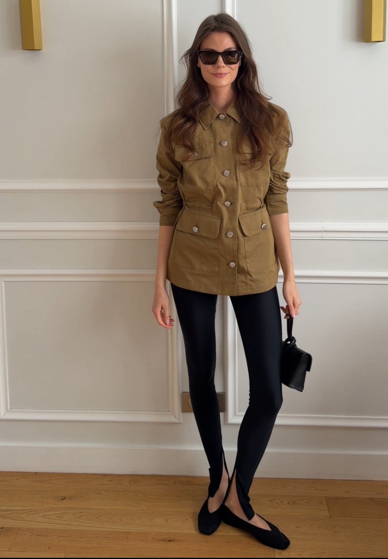 Femme portant une veste marron boutonnée, des leggings noirs avec des fentes à l’avant, des ballerines noires, des lunettes de soleil, et tenant un petit sac à main noir contre un mur blanc.