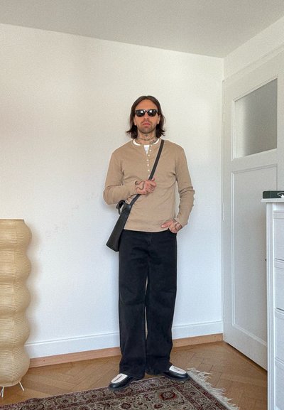 Hombre con gafas de sol, camisa beige de manga larga, pantalones anchos negros, zapatos blancos, bolso cruzado, de pie en el interior cerca de una puerta y una lámpara beige texturizada.