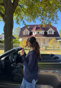 Melna kabrioleta automašīna ar vaļēju jumtu, pelēka interjera. Jauna sieviete tumši zila džempera un džinsu biksēs tur lūpu kosmētikas līdzekli, stāvot pie koka un mājas.