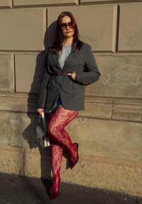 Szary blazer w prążki z paskiem w talii na lekkim swetrze, w połączeniu z czerwonymi koronkowymi rajstopami i błyszczącymi czerwonymi szpilkami, trzymając białą torebkę.
