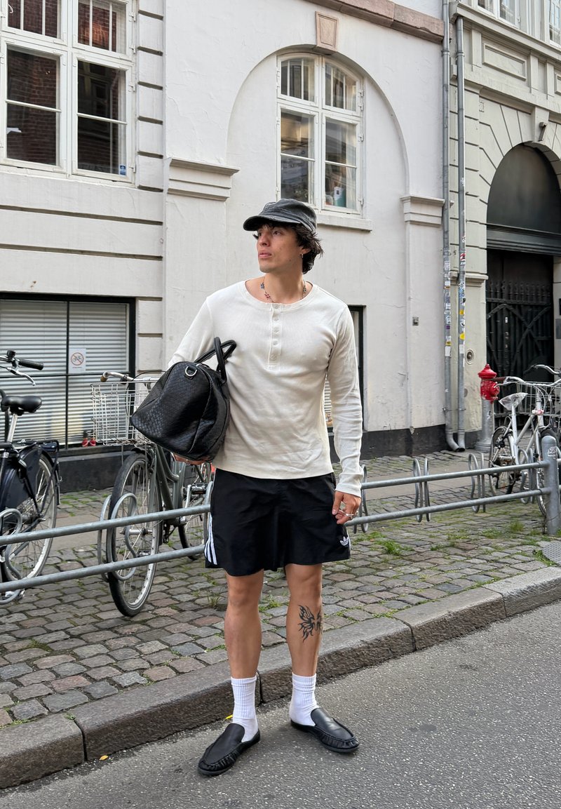 Jeune homme debout dans une rue pavée tenant un sac noir, portant une chemise blanche à manches longues, un short noir, une casquette noire et des chaussures noires.