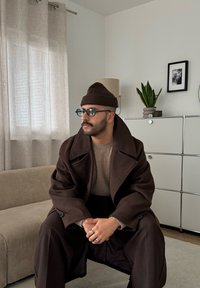 Mężczyzna w brązowym płaszczu, czapce, okularach i pierścionkach siedzący na beżowej kanapie w nowoczesnym salonie z rośliną i oprawionym zdjęciem.