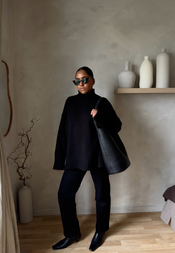 Frau in schwarzem Rollkragenpullover, Hose, Stiefeln und Sonnenbrille hält eine große schwarze Ledertragetasche drinnen neben einem Regal mit Vasen.