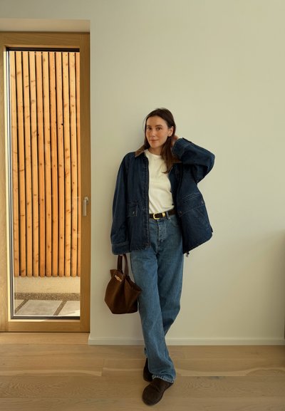 Mujer de pie en el interior junto a una puerta de madera, vistiendo una chaqueta vaquera oscura, camisa blanca, vaqueros azules, sosteniendo un bolso marrón y zapatos oscuros.