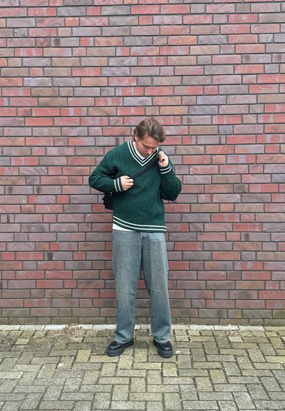 Suéter verde de punto trenzado con rayas blancas, combinado con pantalones grises holgados y zapatos de plataforma negros, frente a un fondo de pared de ladrillo.