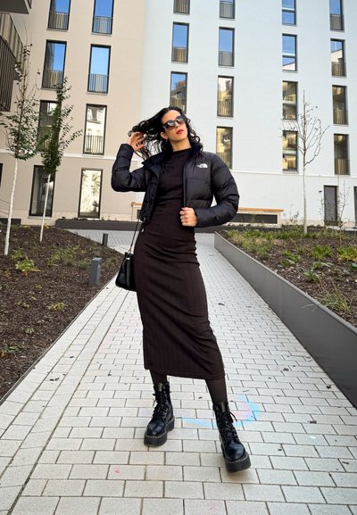 Chaqueta de plumas negra, vestido midi negro ajustado, botas de combate negras con cordones. Pavimento suave con algo de vegetación en un entorno arquitectónico moderno.