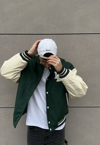 Vihreä ja beige ylioppilastakki, jossa on resoriset hihansuut ja logo. Valkoinen lakki, jossa on teksti "POLO". Päällä valkoinen t-paita ja mustat farkut.