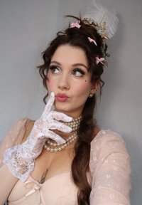 Abito rosa blush con maniche in pizzo, collane di perle a strati, guanti in pizzo e un accessorio per capelli decorativo con fiori e piume.