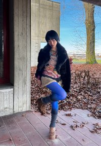 Femme en collants bleus, haut à motifs, veste en fourrure noire, chaussures à imprimé léopard et guêtres grises, posant sur une jambe sur un patio carrelé avec des feuilles d'automne.