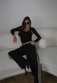 Czarna krótka bluza z długim rękawem, czarne spodnie o szerokich nogawkach z białymi paskami oraz czarne okulary przeciwsłoneczne. Siedzi na jasno szarej, zakrzywionej kanapie z okrągłą poduszką.