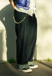 Volné černé džínové kalhoty s ohrnutým lemem, zdobené dekorativním řetízkem z žlutých a černých korálků. Černé tenisky s bílými podrážkami.