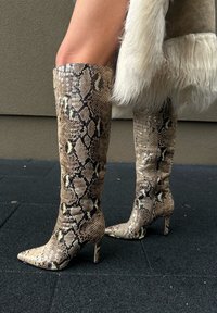 Pernas a usar botas de salto altas com padrão de pele de cobra até ao joelho e um casaco bege com acabamento em pelo branco, de pé sobre um pavimento preto.