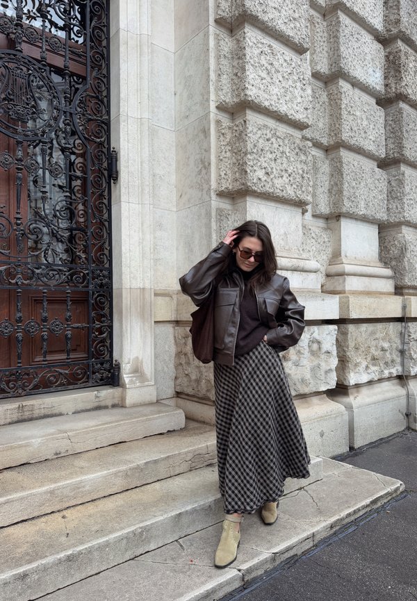 Woman in sunglasses and leather jacket adjusting her hair, standing on stone steps beside an ornate wrought iron gate and a textured building façade.