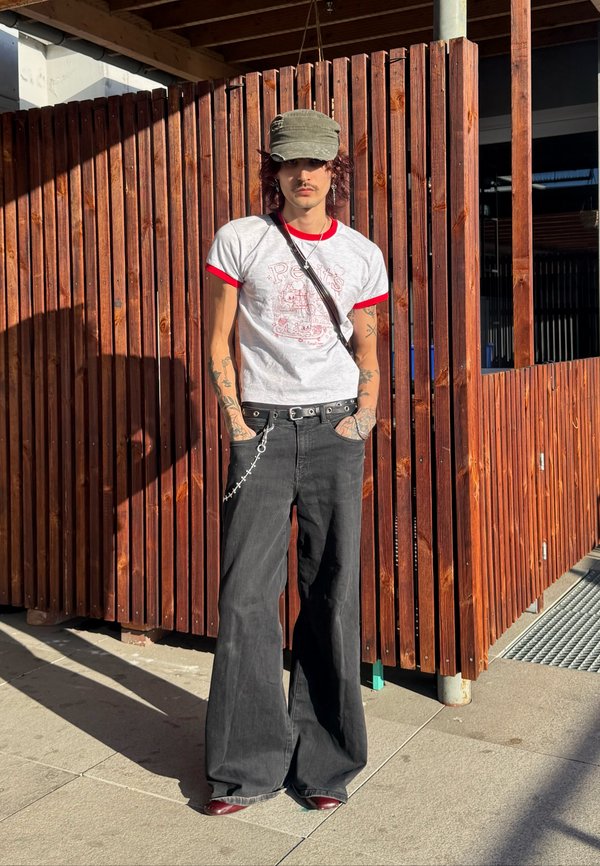 Person mit Tätowierungen, die eine graue Mütze, weißes T-Shirt mit rotem Rand, weite schwarze Hose und Kettengürtel trägt, stehend vor einer Holzgitterwand im Sonnenlicht.