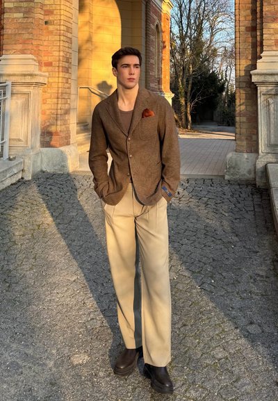 Blazer marrón texturizado con un pañuelo de bolsillo, pantalones de pierna ancha color beige y botas negras, situados sobre un camino de adoquines y arquitectura de ladrillo.