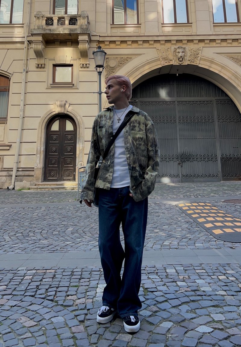 Personne jeune aux cheveux roses portant une veste à motifs, une chemise blanche et un jean large, debout dans une rue pavée près d'un bâtiment historique.