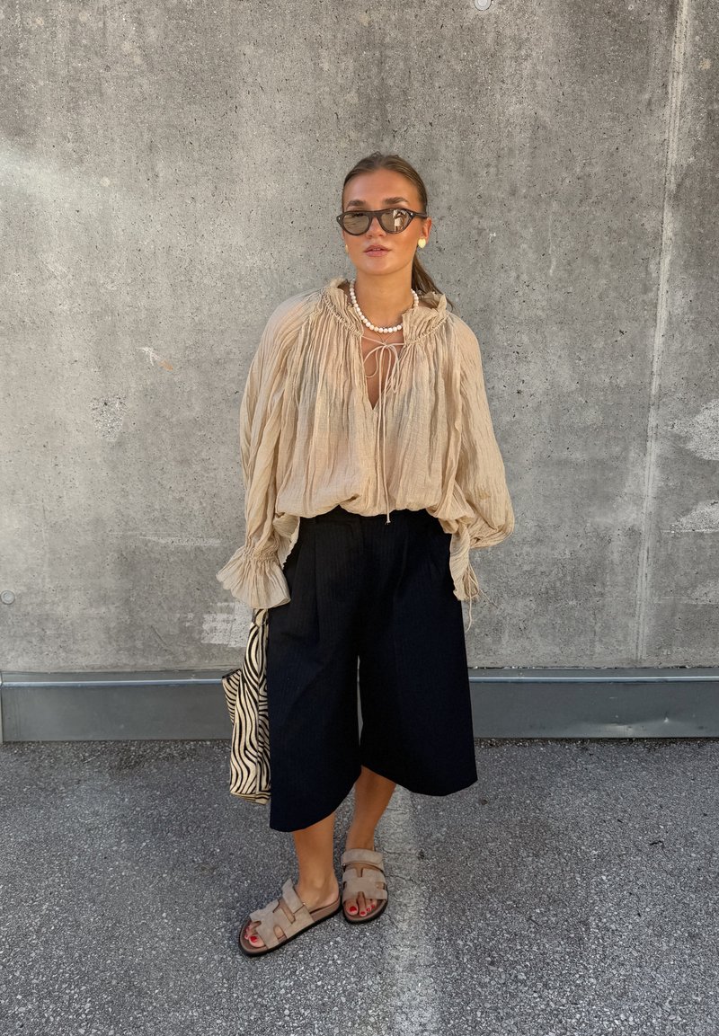Jeune femme portant un chemisier beige ample, un pantalon large noir, des sandales beiges, des lunettes de soleil, et portant un sac à motif zèbre, se tient contre un mur en béton.