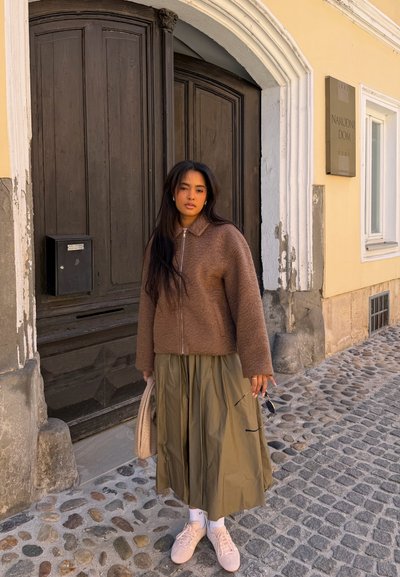 Joven con cabello oscuro y largo que lleva una chaqueta marrón, falda verde oliva, zapatillas blancas y sostiene gafas de sol y un bolso en una calle empedrada.