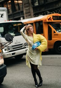 Kollane puffer-tüüpi jope, kollase-valgete triipudega kleit, mustad sukkpüksid, madalad saapad, läbipaistev sinine käekott; linnakeskkond liiklusega taustal.