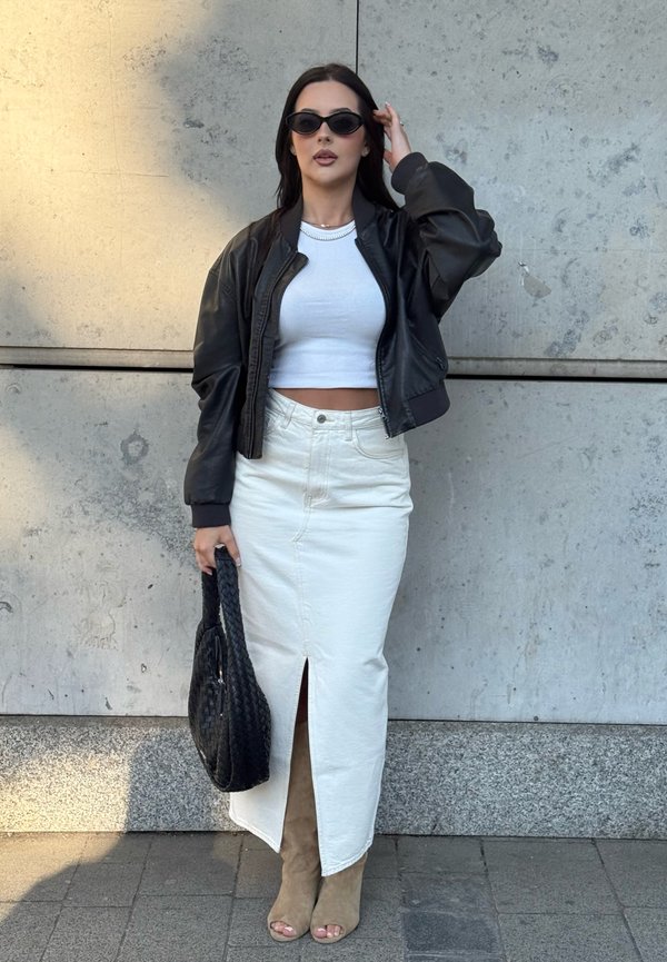 Frau trägt schwarze Lederjacke, weißes Croptop, langen weißen Rock mit Schlitz vorne, beige offene Stiefel, schwarze Tasche und schwarze Sonnenbrille und steht draußen.