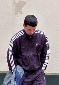 Survêtement violet avec des rayures blanches, veste zippée et pantalon assorti. Tient une chemise à carreaux, debout contre un mur texturé.