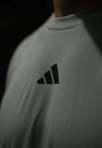 Gros plan sur une personne portant une chemise gris clair texturée avec un logo Adidas noir sur la poitrine.