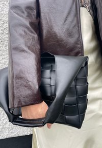 Bolsa de mão em couro trançado preto com uma alça grossa e macia. Combinada com um casaco bordeaux e um vestido de seda de cor clara, destacando os contrastes de textura.