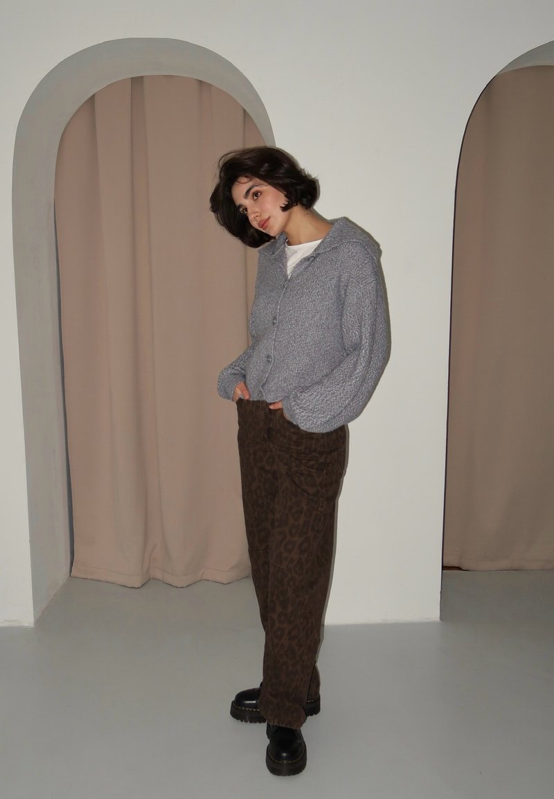 Γκρι πλεκτό πουλόβερ με κουκούλα, κλείσιμο με κουμπιά, φορεμένο πάνω από ένα λευκό πουκάμισο. Καφέ παντελόνι με λεοπάρ σχέδιο και μαύρες πλατφόρμες ολοκληρώνουν την εμφάνιση.