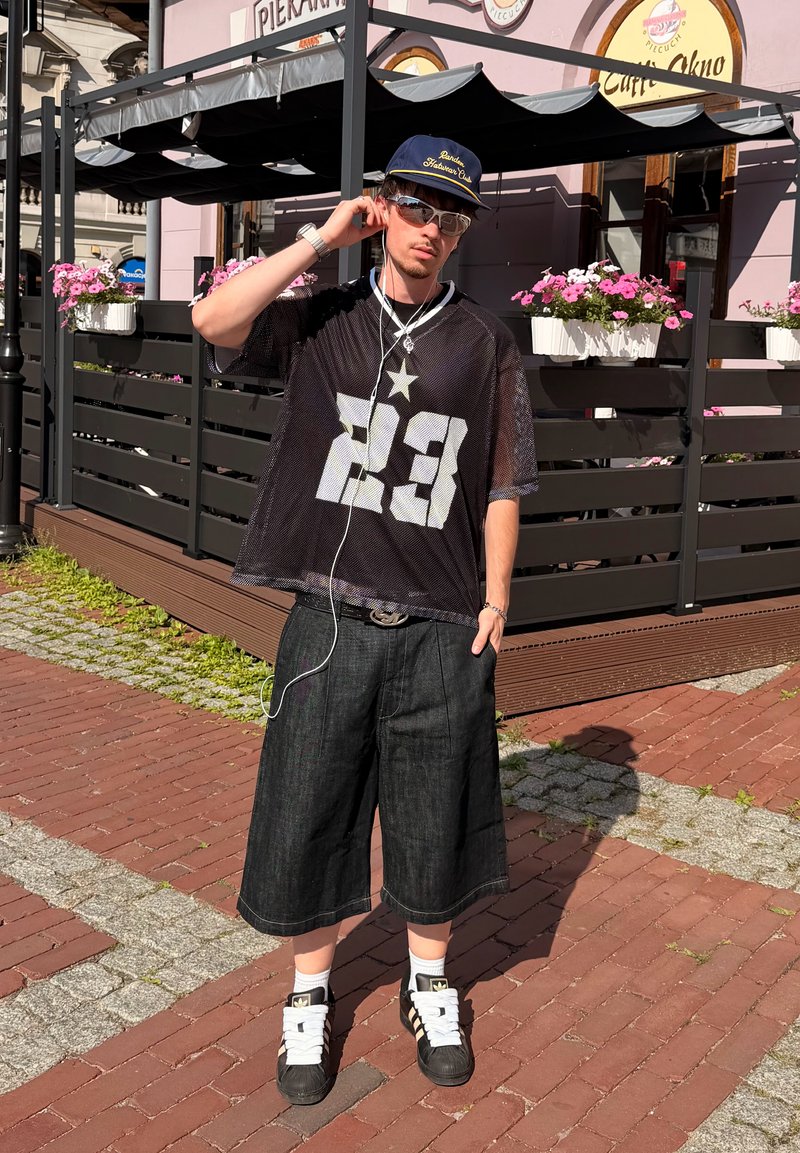 Jeune homme portant un t-shirt en mesh noir avec le numéro 23, un short en jean large, des baskets, des lunettes de soleil et une casquette, debout sur un trottoir en briques.