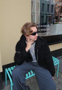 Asmuo sėdi ant turkio spalvos metalinės suoliuko, dėvi akinius nuo saulės, juodą striukę, kamufliažinę palaidinę ir pilkus plačiomis kojomis kelnes.