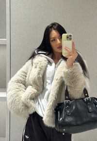 Krycí kabát z umělé kožešiny ve světle béžové barvě, se zipem, spojený se šedou mikinou, černými sportovními kalhotami a strukturovanou černou kabelkou se stříbrnými doplňky.