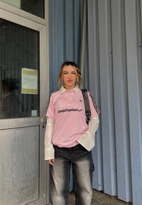 Roza adidas majica s črnim logotipom, oblečena čez belo dolgo majico z dolgimi rokavi. Črne kavbojke z zbledelim videzom. Stoji ob kovinskih vratih.