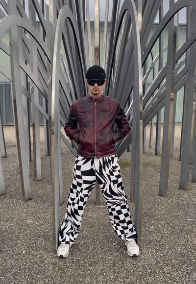 Chaqueta de cuero marrón con mangas negras, pantalones a cuadros en blanco y negro, gorro negro y gafas de sol, de pie entre estructuras metálicas.