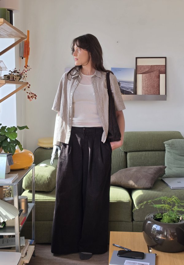 Mujer con antebrazo izquierdo protésico está de pie en una sala de estar, llevando una camisa de cuadros, camiseta blanca, pantalones negros y llevando una bolsa negra.