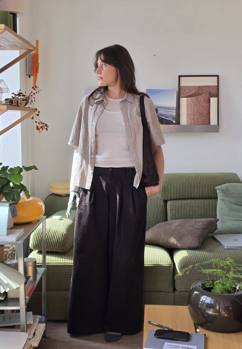 Femme avec un avant-bras gauche prothétique debout dans un salon, portant une chemise à carreaux, un haut blanc, un pantalon noir, et portant un sac cabas noir.