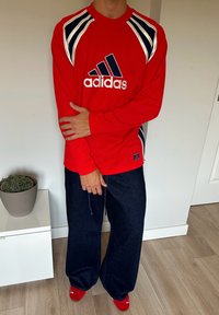 Rød langermet Adidas-trøye med mesh-lignende tekstur, med svarte og hvite detaljer på skuldrene og logoen. Brukt med mørke denim-bukser.